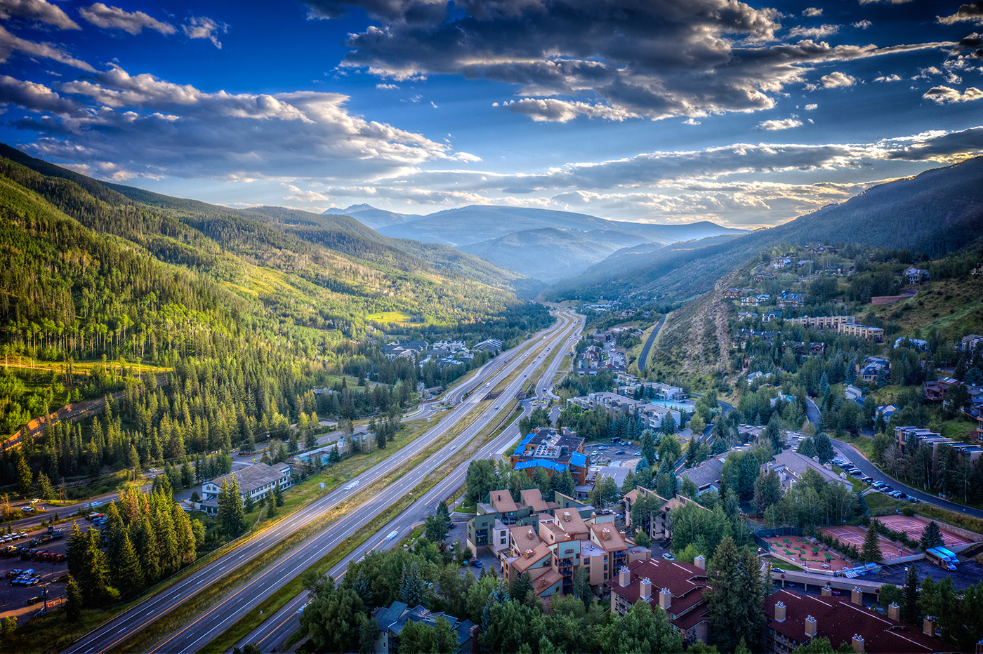 Drone shot of Vail Valley, Colorado, in the spring with evergreen trees peppered throughout the image, between buildings, and all along the hills raising up to mountains on a beautiful afternoon, with the sun peaking through the clouds in a blue sky